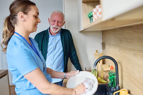 A caregiver cleans dishes at a kitchen sink with an elderly person observing.